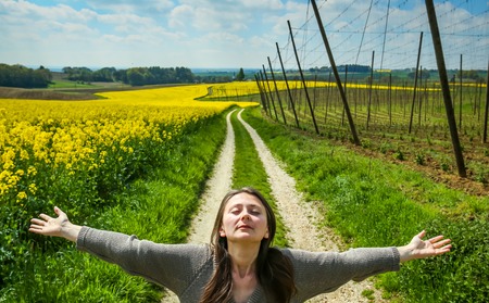 A woman spreading her arms standing on a pathway leading through the yellow flowering rapeseed and hop fields in spring.の写真素材
