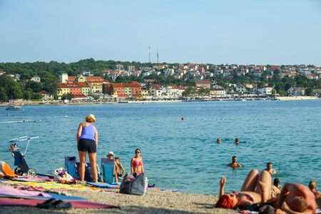 NJIVICE, CROATIA - JUNE 24, 2017 : People bathing and sunbathing on sea beach with town panorama in...