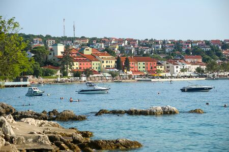 NJIVICE, CROATIA - JUNE 24, 2017 : People swimming in the sea with the anchored boats and colorful architecture of hotel Jadran in the background in Njivice, island Krk, Croatia.のeditorial素材