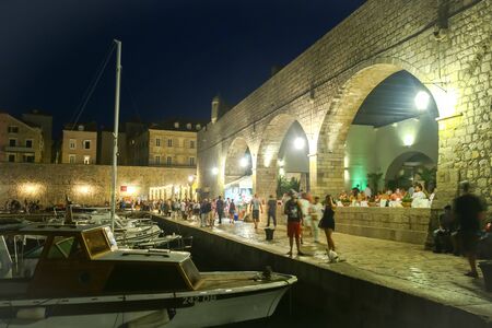 DUBROVNIK, CRATIA - JULY 22, 2017 : People walking on the seafront next to a restaurant with lined up moored boats in the city harbor at evening in Dubrovnik, Croatia.のeditorial素材