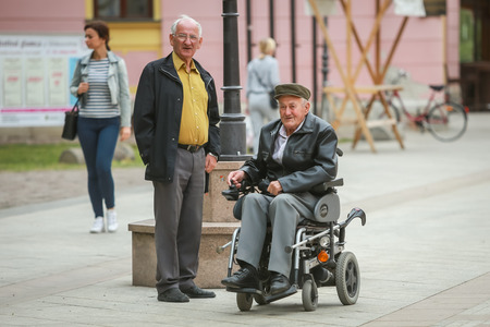 VINKOVCI, CROATIA - MAY 14, 2018 : Close up of two senior men while one is in the wheelchairs and driving in the street of Vinkovci, Croatia.のeditorial素材