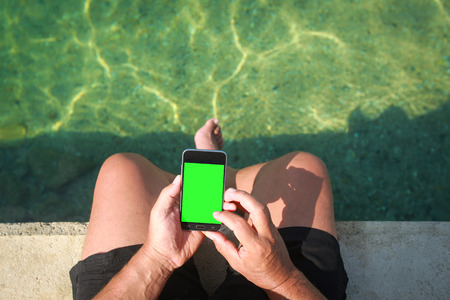 A man holding smartphone while sitting on the seaside pier, green screen cut out.の写真素材