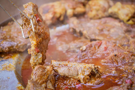 Detail of a human hand turning a pork steak meat cooked in vegetable sauce together with sausages and chicken meat on a barbeque fire in a traditional croatian kotlovina dish.の写真素材