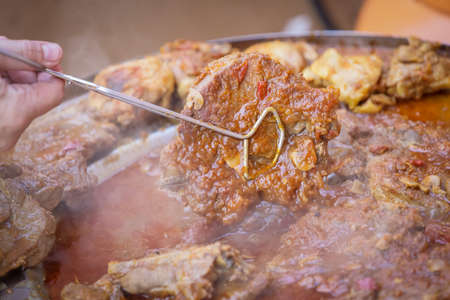 Detail of a human hand turning a pork steak meat cooked in vegetable sauce together with sausages and chicken meat on a barbeque fire in a traditional croatian kotlovina dish.の写真素材