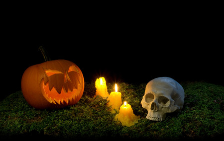 Halloween pumpkin, human skull and candles glowing in the dark on a forest moss. Decoration of the party emerges from the dark and illuminates the green vegetation.の写真素材
