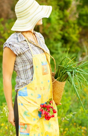 Happy woman gardener working on field, young female holding basket, girl growing organic green vegetables and fruits, summer garden, rural leisure outdoor, lady farmer, radish and onion harvest seasonの写真素材