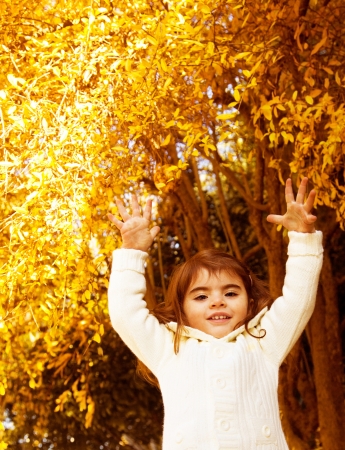 Image of adorable small girl play game on backyard in fall, candid cheerful kid in warm white sweater enjoying beautiful autumn season with raised hands up, golden autumnal weatherの写真素材