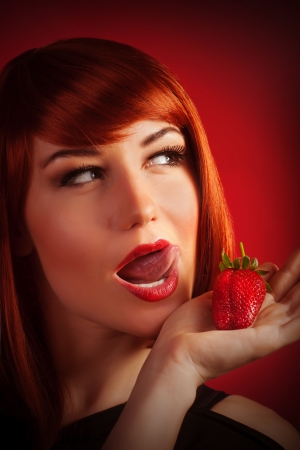 Picture of attractive female with open mouth isolated on red background, sexy red hair woman eating fresh strawberry, healthy lifestyle, fruity diet, Valentine day, pleasure and enjoying conceptの写真素材