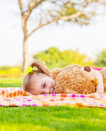 Adorable child resting on backyard in spring time, baby girl laying down on green field with teddy bear, having fun outdoors, happy childhood conceptの写真素材