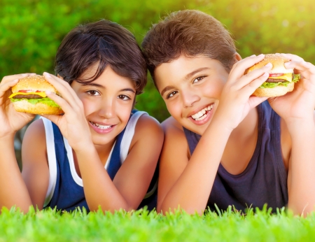 Closeup portrait of two happy boys eating big tasty fatty burgers outdoors, lying down on green field and enjoying sandwich with cheese, meat and vegetablesの写真素材