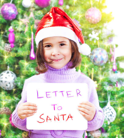 Closeup on cute little girl holding in hands letter to Santa Claus, wearing red festive hat, beautiful decorated Christmas tree, winter holidays conceptの写真素材