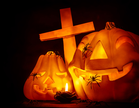 Closeup on glowing carved pumpkin with creepy spiders on graveyard, mysterious cross on dark Halloween nightの写真素材