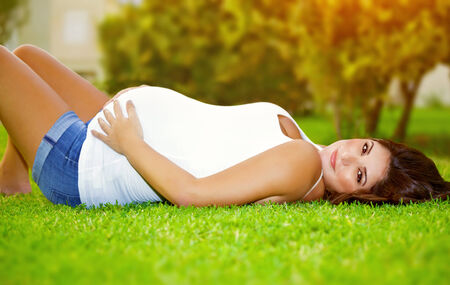 Woman awaiting for a baby, cute pregnant female lying down on fresh green grass in the garden, sunny...