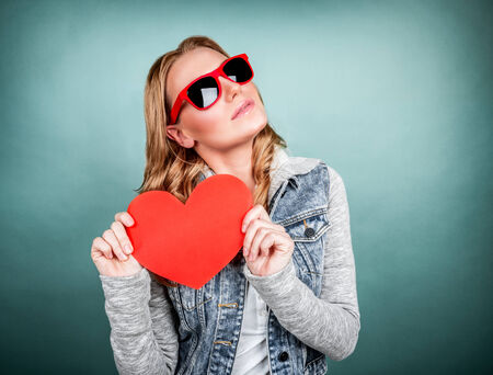 Cute girl with red heart isolated on blue background, wearing stylish sunglasses, greeting card for Valentine day, modern love concepの写真素材
