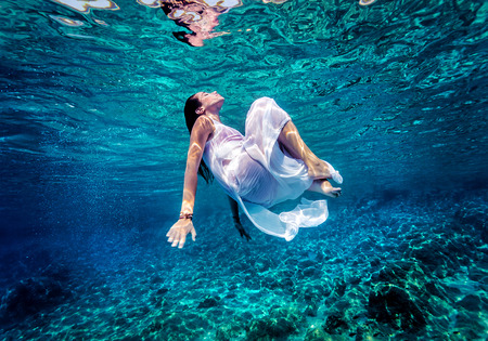Gorgeous female dancing underwater, wearing long white fashion dress, summer activity, relaxation in blue transparent sea, enjoyment and refreshment conceptの写真素材
