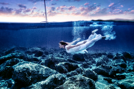 Young woman diving in sunset time, enjoying swimming underwater, wearing long white dress, luxury summer vacation, freedom and pleasure conceptの写真素材