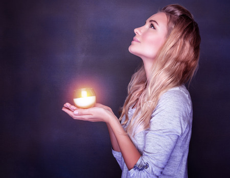 Portrait of beautiful blond girl with glowing candle in hands on dark background, looking up and praying with hope, traditional Christian holiday, Christmas time conceptの写真素材