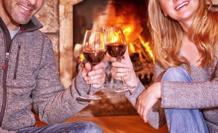 Happy young couple sitting near fireplace and drinking wine, enjoying romantic date, celebrating Valentines day in a cozy winter houseの写真素材