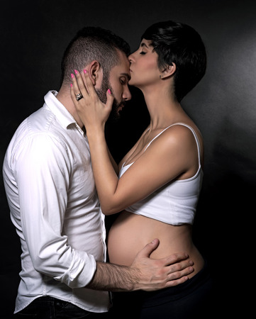 Side view photo of beautiful gentle pregnant woman kissing her loving husband over black background, young family and love conceptの写真素材