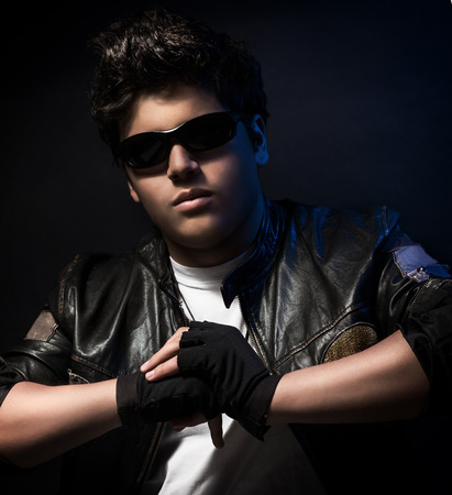 Portrait of beautiful teen boy wearing stylish sunglasses, leather jacket and special sportive gloves over blue background, fashion accessories for teenagersの写真素材