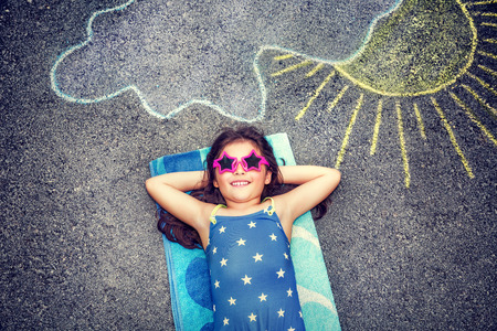 Happy little girl wearing swimsuit and stylish sunglasses lying down on the asphalt near picture of...
