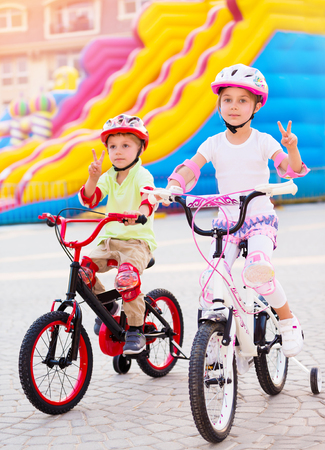 Happy friends on the bicycles, brother with sister having fun in amusement park, gesturing peace by hands, playing game outdoors, enjoying friendship and summer holidaysの写真素材