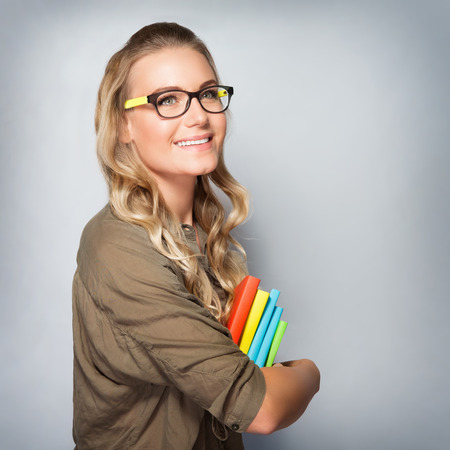 Portrait of cute blond student girl over gray background, wearing glasses and holding in hands colorful books, back to schoolの写真素材