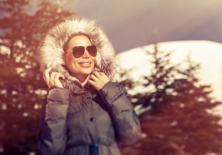 Portrait of beautiful woman having fun in the winter park, wearing sunglasses and coat with fur hood...