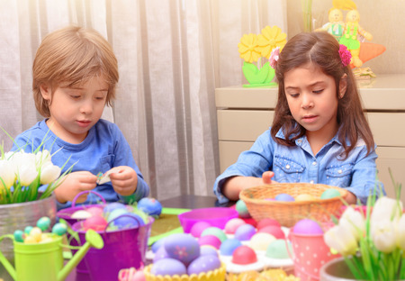 Portrait of little brother and sister painting traditional Easter eggs in different colors at home,...