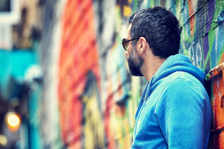 Profile of a stylish handsome guy wearing sunglasses and casual clothing standing near beautiful colorful city wall background, looking away, urban lifestyleの写真素材