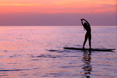 Silhouette of a girl practicing yoga, balancing on the surf board in the sea over beautiful purple...