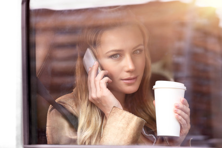 Portrait of a beautiful business woman sitting in the car with cup of coffee, talking on the phone...