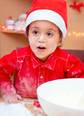 Portrait of a cute little boy soiled in flour, wearing red Santa hat making Christmas cookies at home, happy festive time, preparation for winter holidaysの写真素材