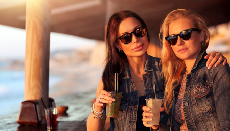 Portrait of a two beautiful girls spending leisure time in the outdoor cafe with drinks, models wearing stylish sunglasses, enjoying bright sunny dayの写真素材
