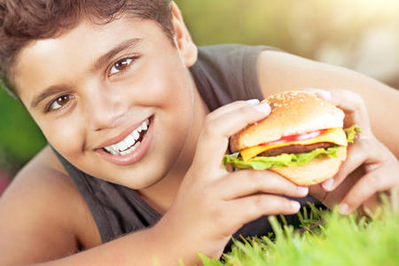Closeup portrait of a cute happy teen boy lying down on green grass and eating delicious burger, enjoying tasty lunch on sunny day, happy time at summer campの写真素材