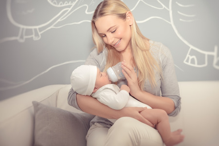 Cute mother feeding her adorable newborn baby in the beautiful cozy kid's room at home, special milk formula for infants, enjoying parenthood, happy family lifeの写真素材