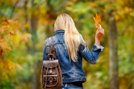 Rear view of a blond girl walking in the forest, holding in hand dry tree leaf, having fun in autumn...