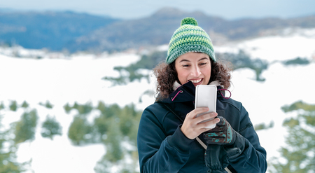 Cute cheerful woman using mobile phone in the mountains, receiving funny messages, having fun outdoors, with pleasure spending winter vacation in the snowy mountainsの写真素材