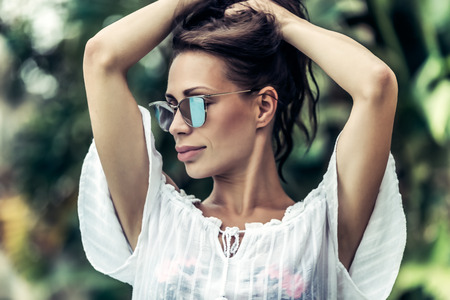 Portrait of a gorgeous female wearing stylish sunglasses posing over exotic plants background, photoshoot on the luxury beach resort, enjoying summer vacationの写真素材