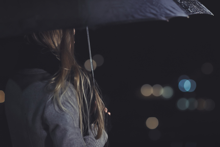Female outdoors in rainy night, rear view of the elegant woman under black umbrella looking on the city lights, city life, loneliness and melancholy conceptの写真素材