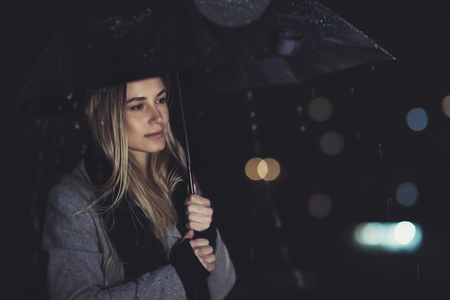 Fashion portrait of beautiful gorgeous woman standing with umbrella underthe rain over blury night...