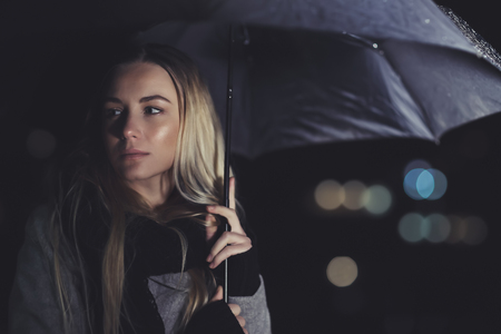 Portrait of a beautiful sad woman waiting for someone at night, standing with umbrella under rain over dark night background with bokeh lights behindの写真素材