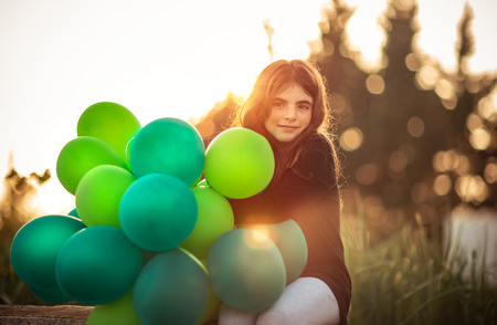 Portrait of a cute little girl sitting in the park with pile of green air balloons outdoors in mild evening sun light, celebrating birthday, preparation to happy holidayの写真素材