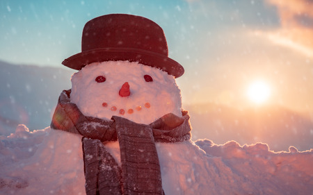 Photo of a cute snowman dressed in a stylish hat and scarf in a snowy frosty evening on a sunset background, happy winter holidays traditionの写真素材