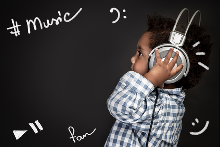 Cute little african american boy wearing earphones, listening and recording baby songs,  portrait over blackboard background, happy little singerの写真素材