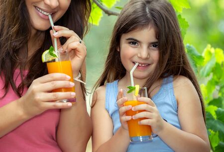 Portrait of a joyful mother and daughter drinking tasty fresh orange juice outdoors, having breakfast in the gardenの写真素材