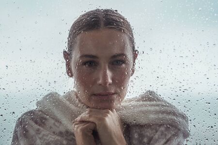 Portrait of a sad woman looking from outdoors into the house through a window covered with...
