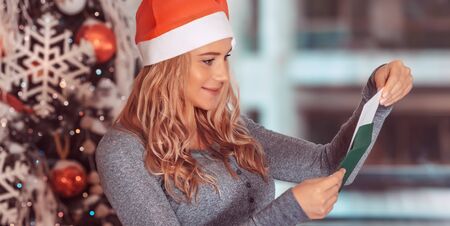 Portrait of a nice woman near Christmas tree with pleasure reading festive postcard with best...