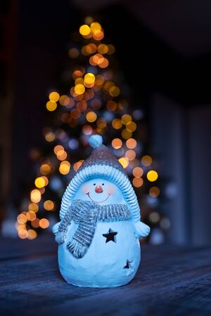 Little decorative snowman toy on the table over glowing Christmas tree background, happy winter holidays, traditional New Year symbolの写真素材