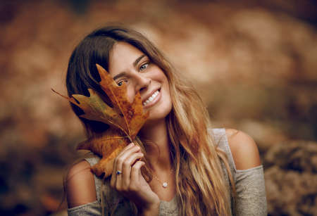 Portrait of a Beautiful Girl Having Fun in Autumn Forest. Holding in Hand Dry Maple Leaf. Beauty of Fall Season.の写真素材